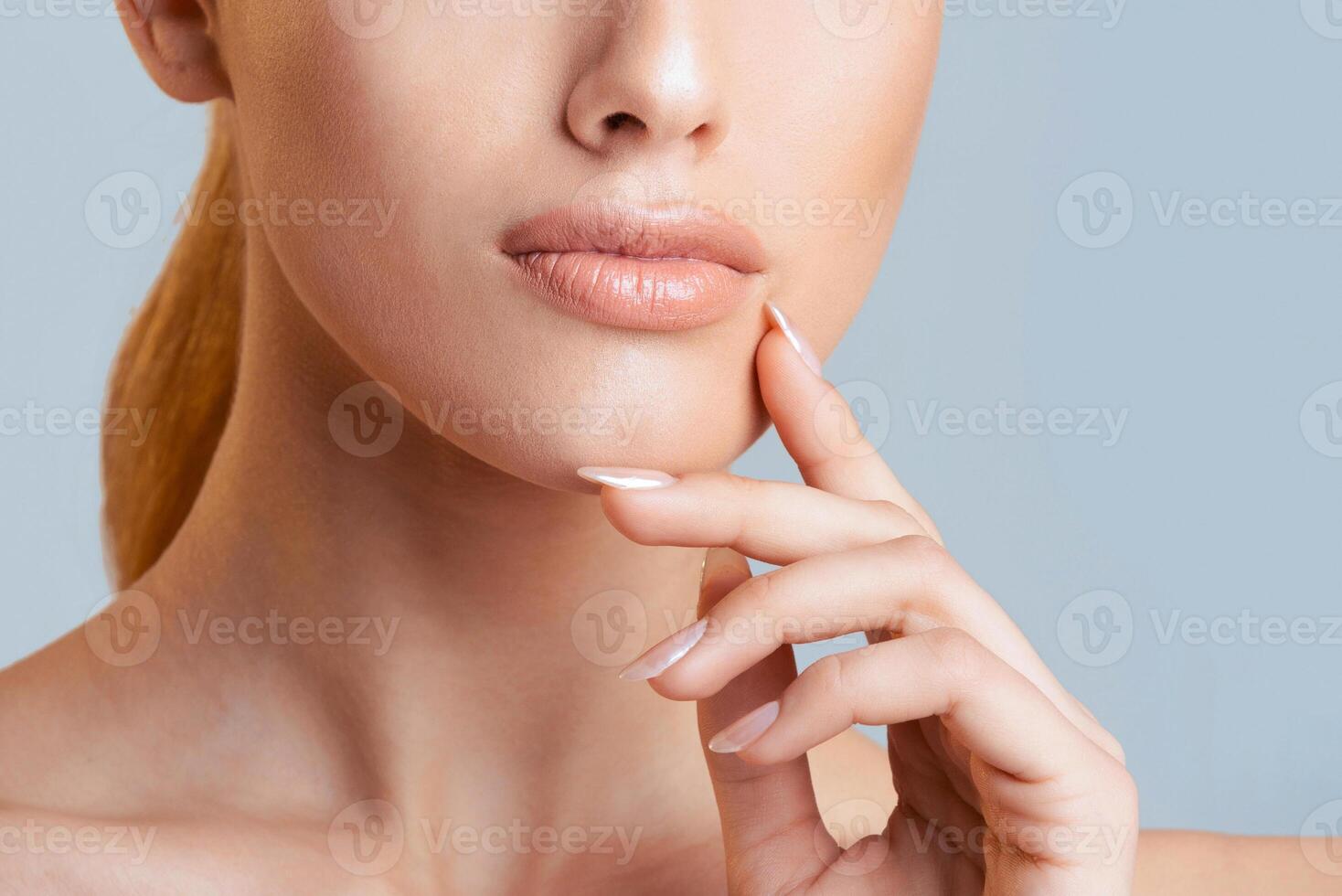 Cropped of woman face with her hand resting on her chin. The woman has light skin, and her lips are slightly parted. Her nails are polished with a light pink color. The background is a soft blue. photo