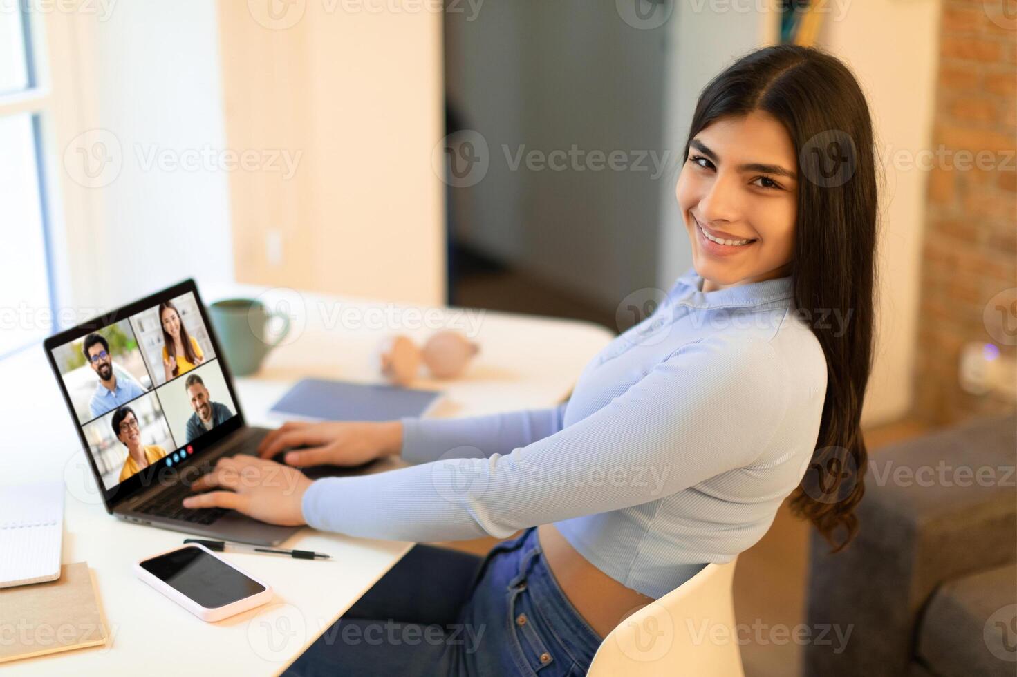 Smiling woman sits at a desk in a home office, smiling and looking at the camera while participating in a conference on her laptop. The conference window shows several faces photo