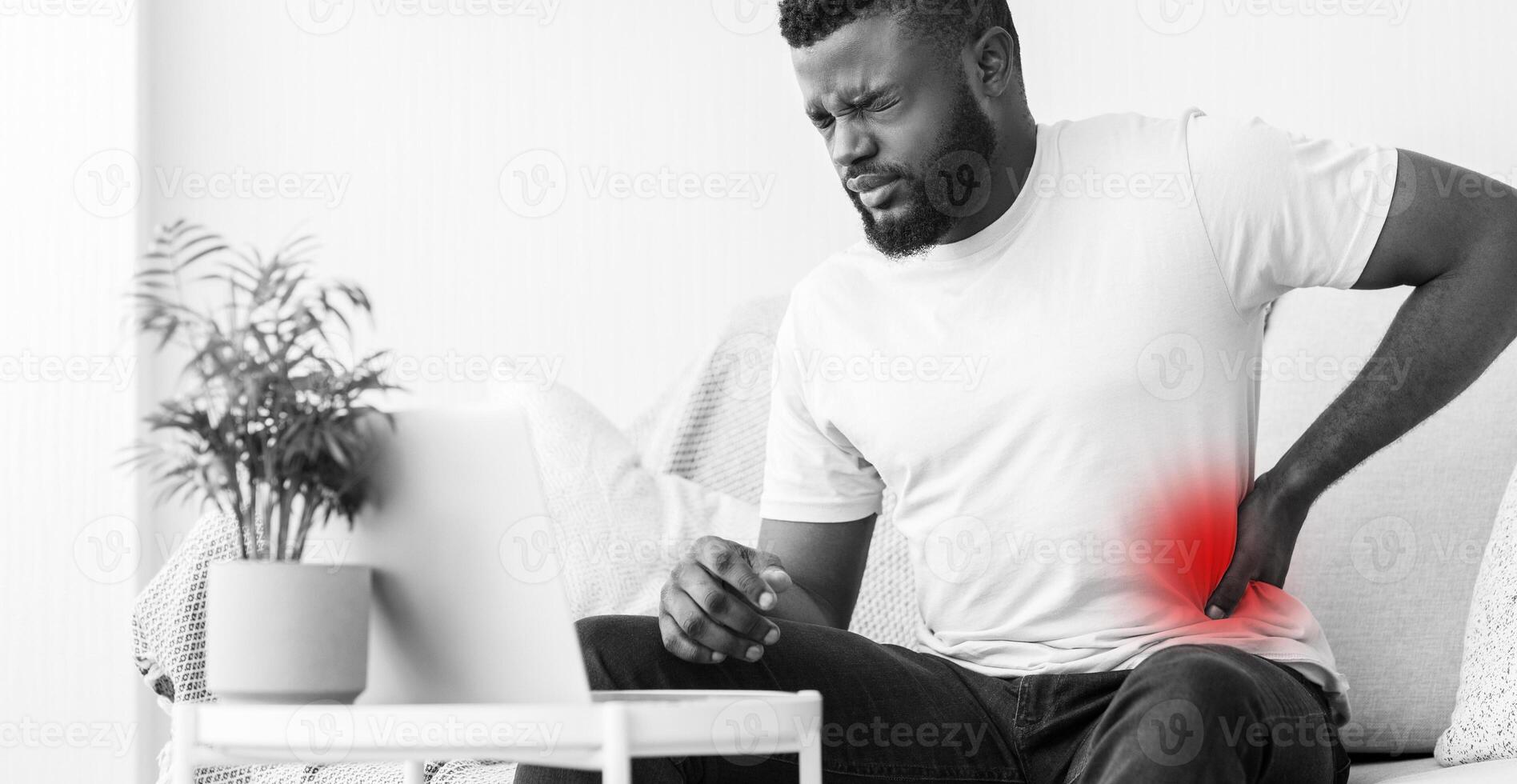 A black and white photo of a man sitting on a couch at home, holding his lower back in pain. He appears to be in discomfort, possibly due to prolonged sitting.