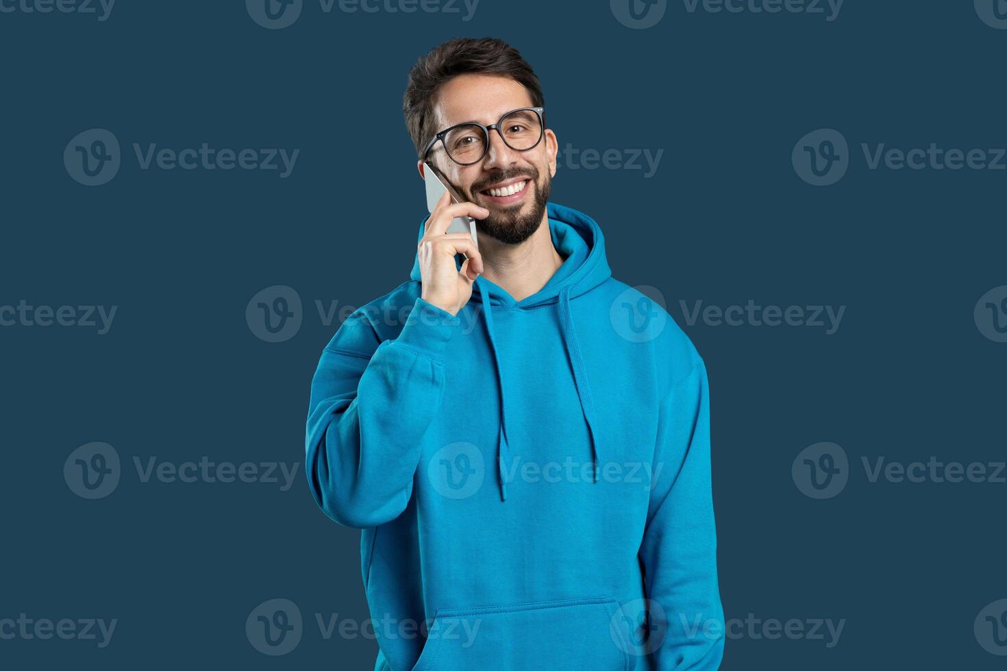 A young man dressed in a blue hoodie smiles bright while speaking on his phone. The warm expression on his face suggests he is engaged in a pleasant conversation photo