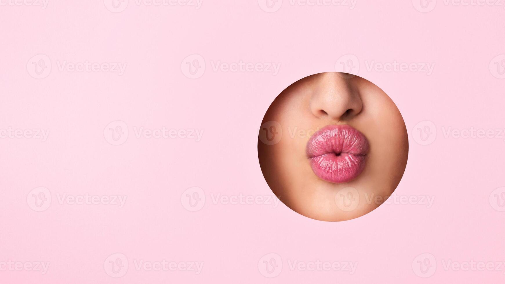 A close-up view of a woman lips protruding out of a hole against a vibrant pink background. The lips appear to be puckered or in a kissing gesture. photo
