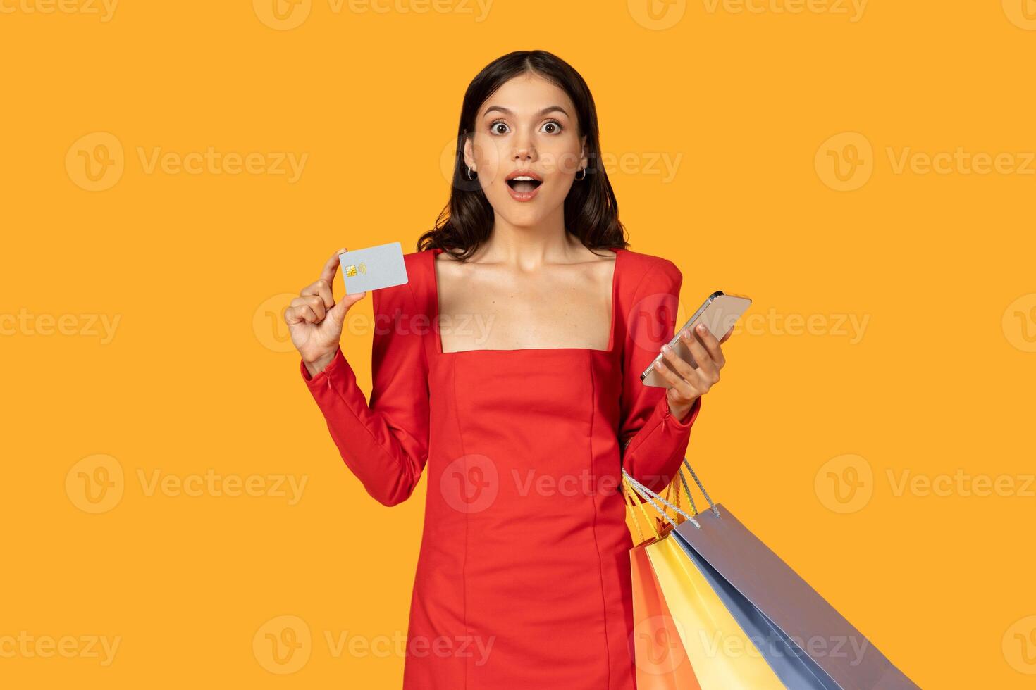 A young woman in a red dress is holding a credit card and smartphone in front of a yellow background. She is looking at the camera with a shocked expression on her face. photo