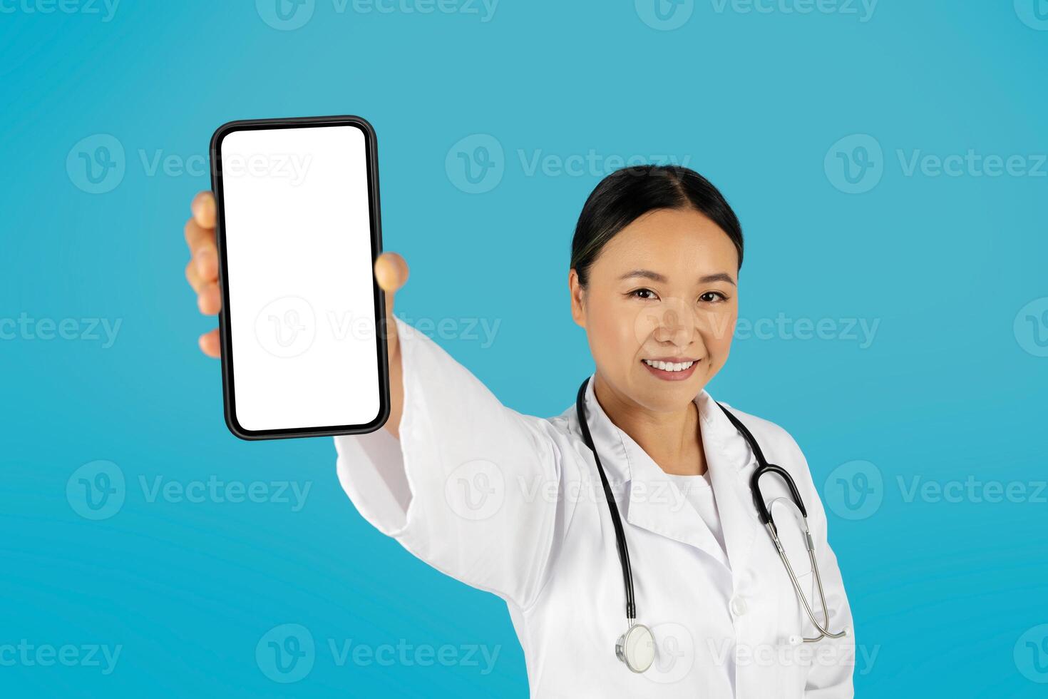 Asian woman doctor wearing a white coat and stethoscope smiles at the camera while holding a smartphone with a blank screen in her right hand. The background is a bright blue. photo