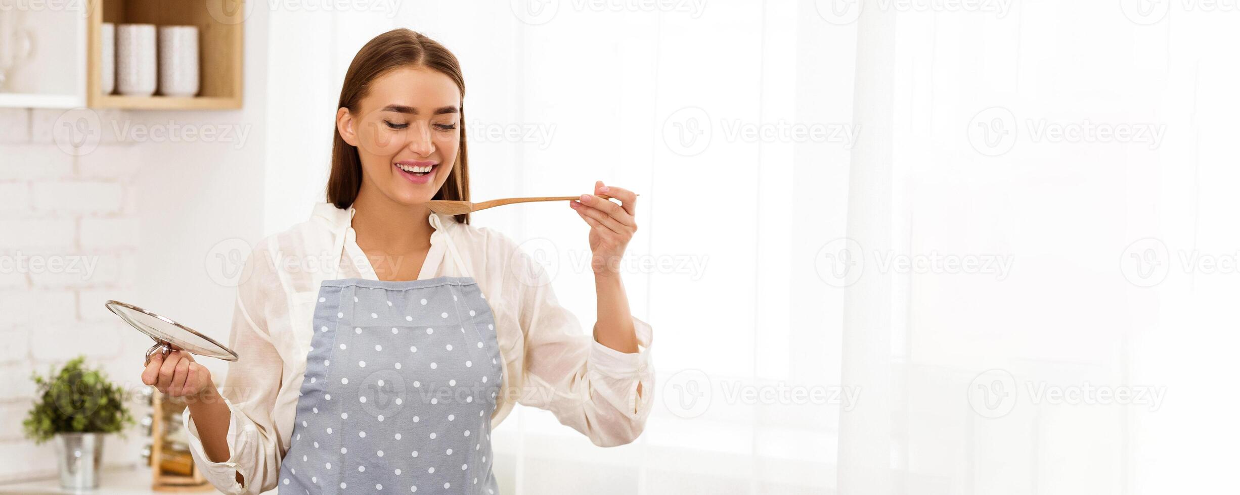 A woman wearing an apron in a kitchen, holding a cooking utensil and tasting food with a spoon. photo