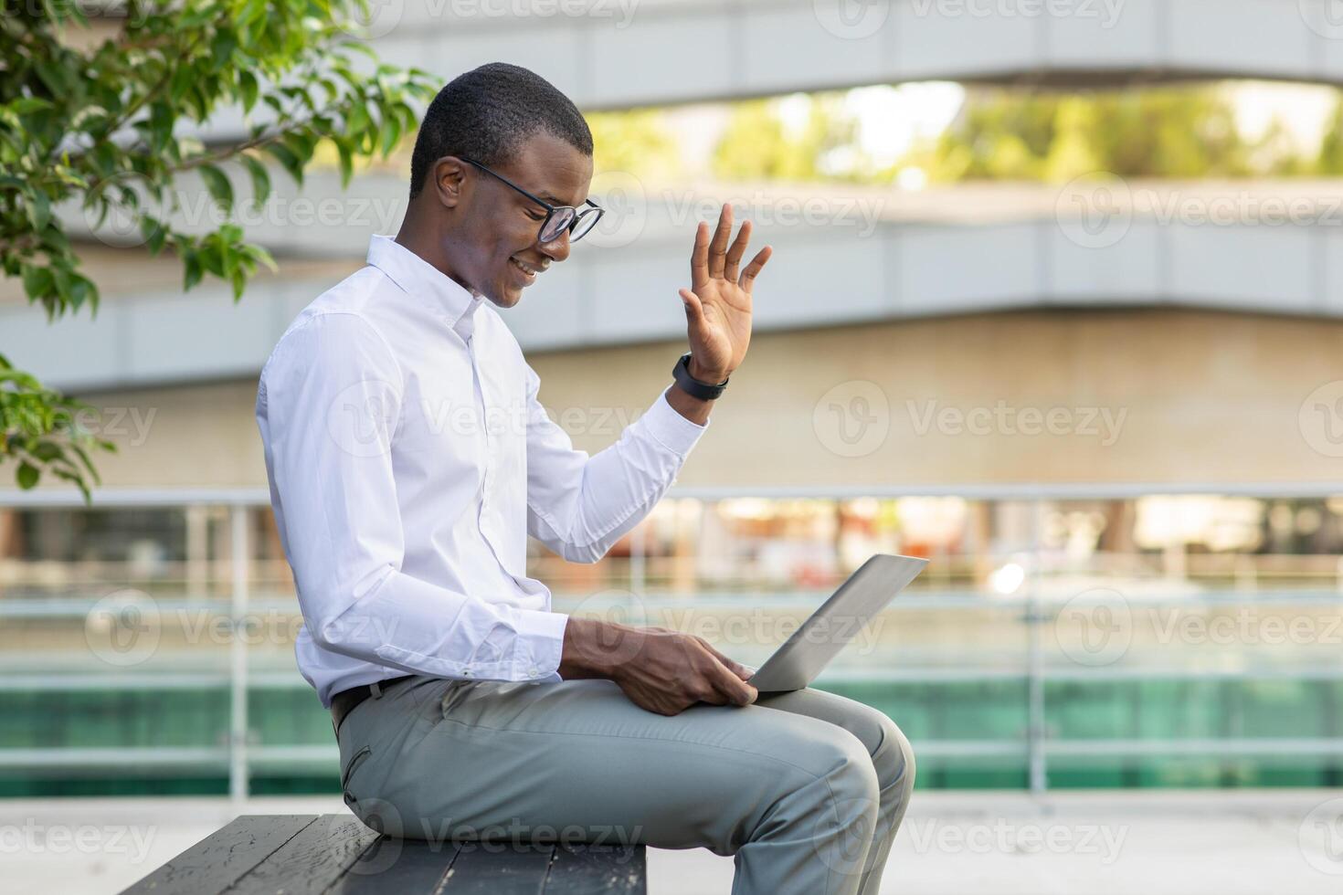 A young African American professional wearing glasses is happily participating in a virtual meeting. He sits on a wooden bench in an outdoor urban area, waving towards the screen of his laptop. photo