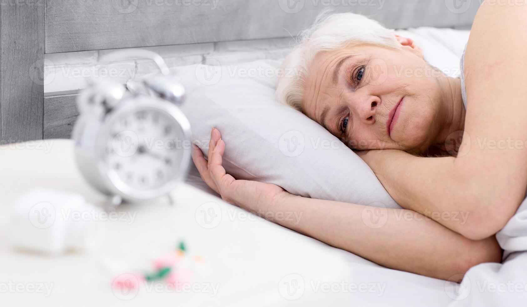 A woman is laying in bed, with her eyes open, next to an alarm clock set on the bedside table. The clock displays a time indicating the morning hour. photo