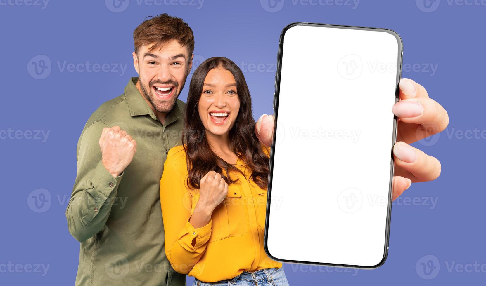 A young man and woman are excitedly celebrating while holding a smartphone with a blank screen in front of them against a blue background. photo