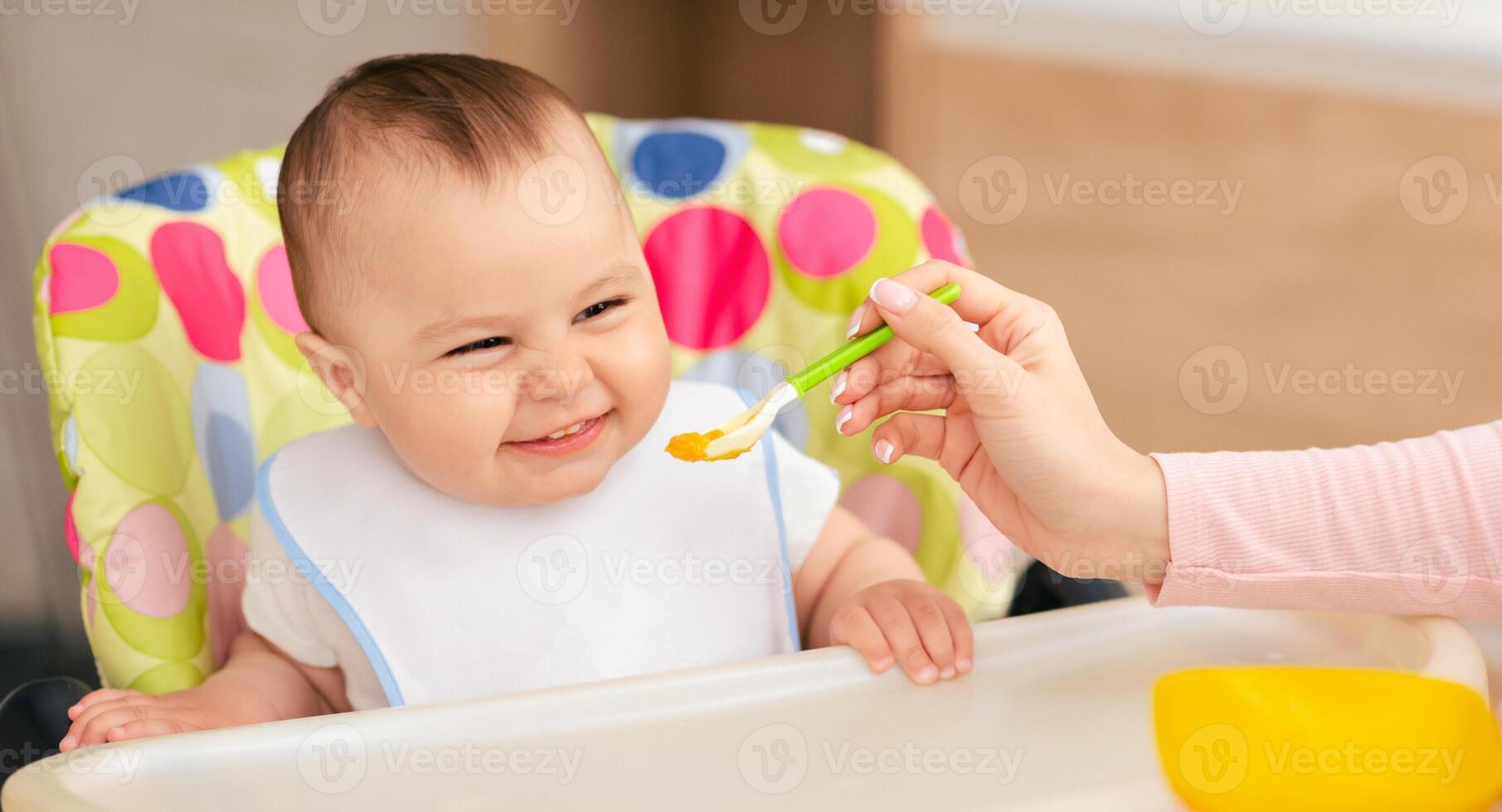 A woman is sitting and feeding a baby with a spoon. The woman hand is holding the spoon, while the baby is looking at the spoon. The baby seems to be enjoying the feeding process. photo