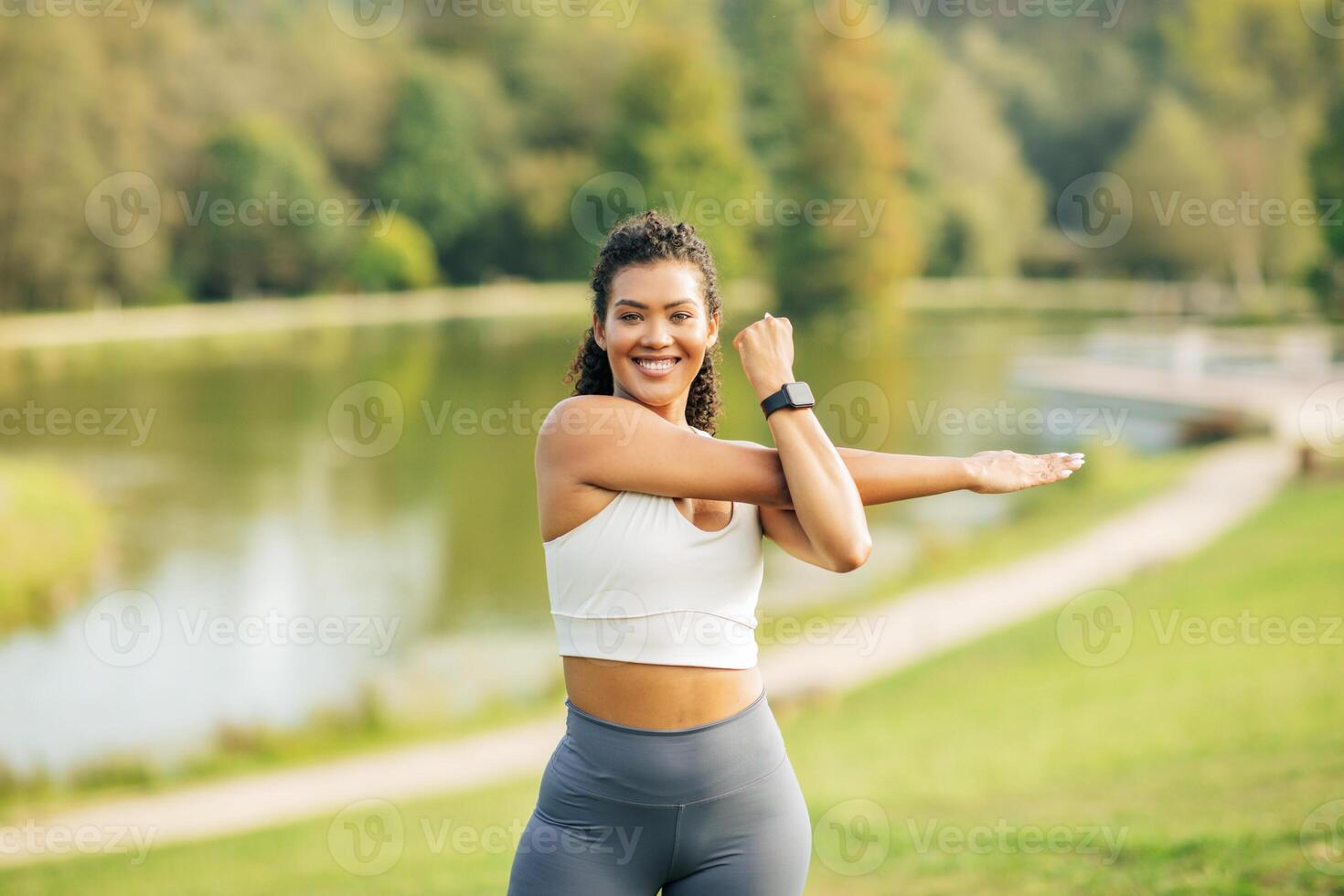 A young woman performs an arm stretch while standing in a lush park beside a serene lake. She is dressed in athletic wear, smiling, and appears to be enjoying her exercise routine on sunny day. photo