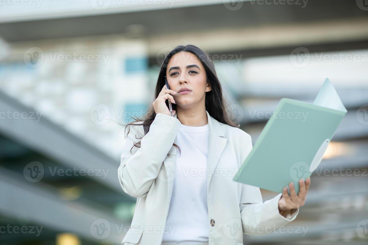A woman in a light suit engages in a phone conversation while holding a file, standing outside a modern building. photo