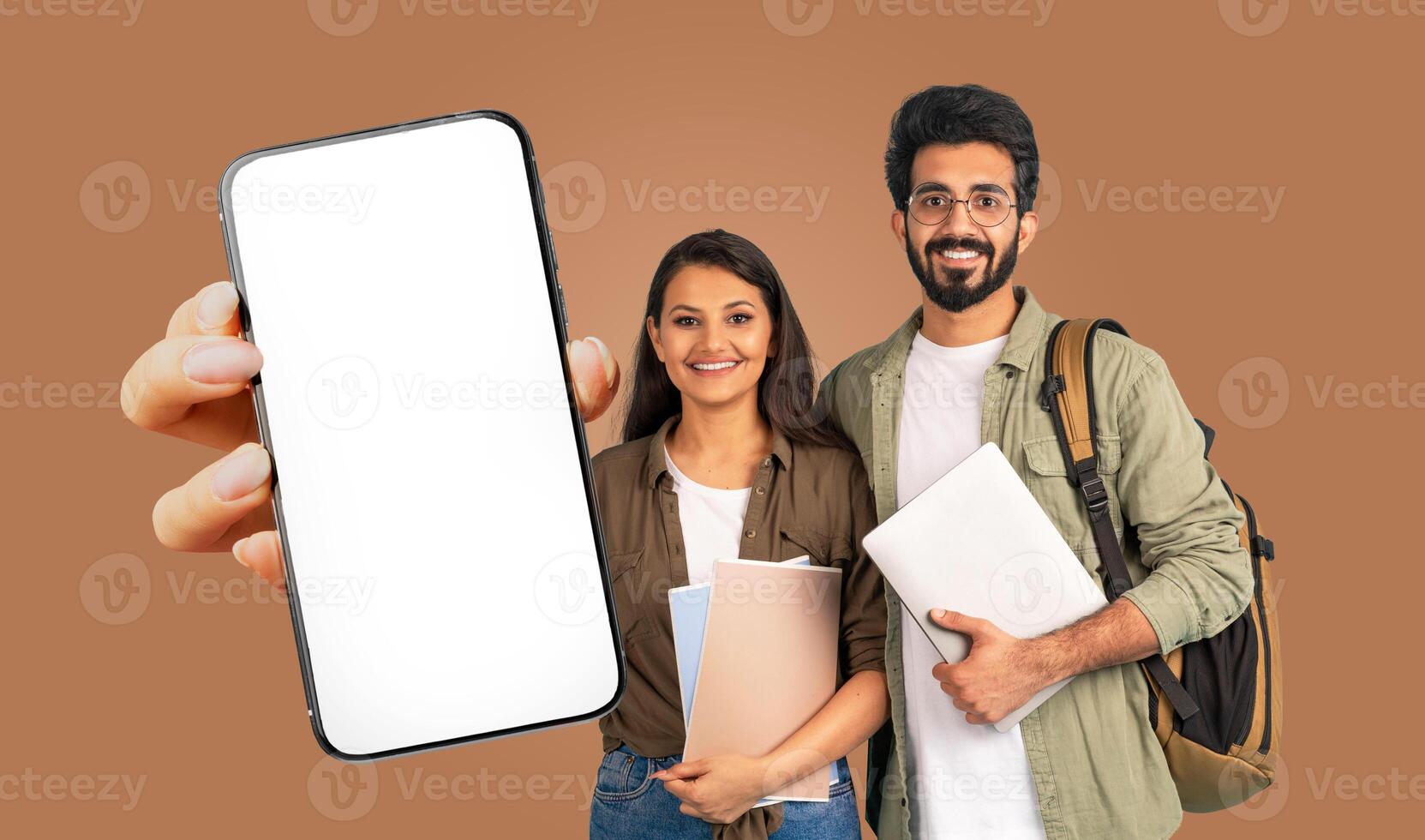 Two smiling Indian students stand in front of a brown background. They are holding books and a smartphone with a blank screen photo