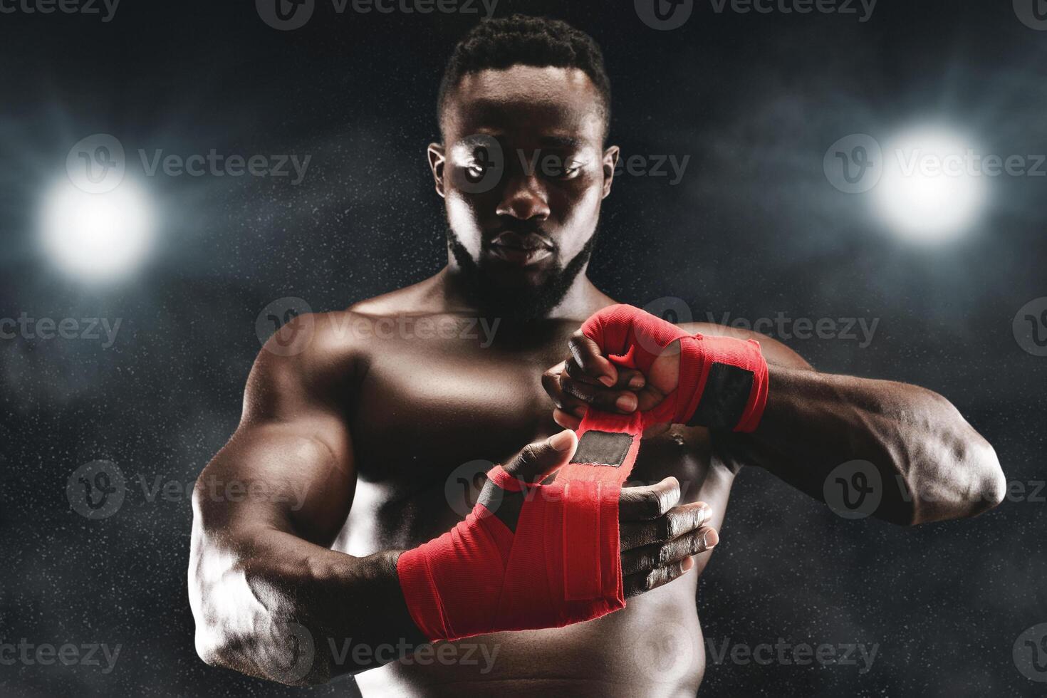 A muscular boxer with a serious expression wraps his hands with red tape in a dimly lit boxing ring. photo