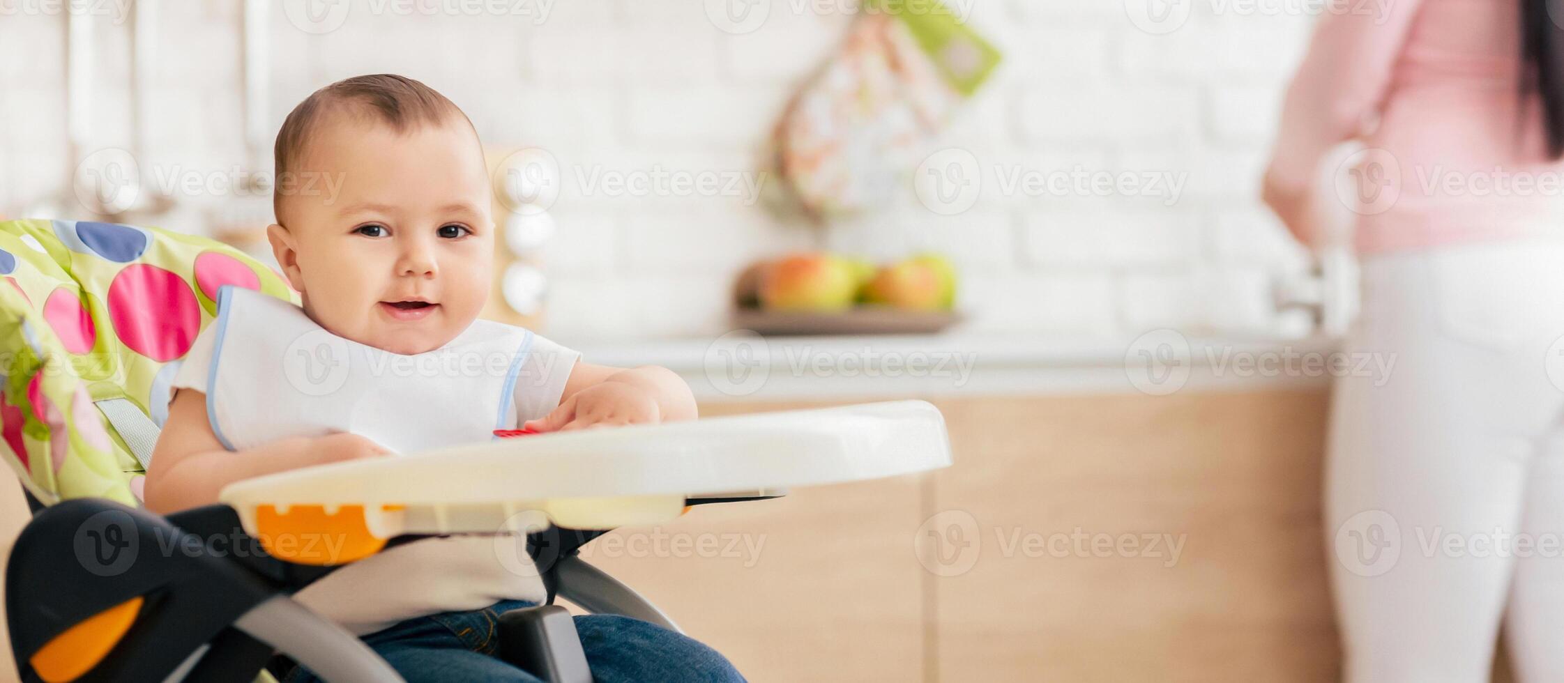 A baby boy is enjoying his meal in a child chair in the kitchen. The setting is warm and inviting, capturing a moment of contentment and family routine. photo