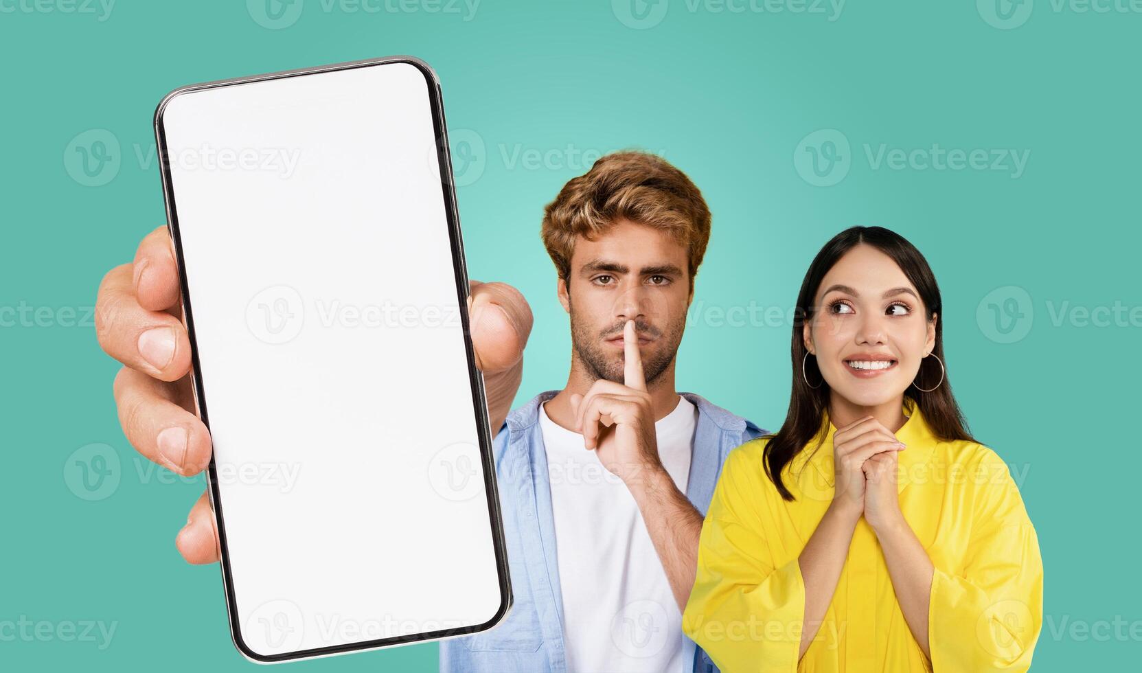 Smartphone with a white blank screen in front of a man and woman. The man is holding his finger to his lips, while the woman smiles and has her hands clasped in front of her. photo