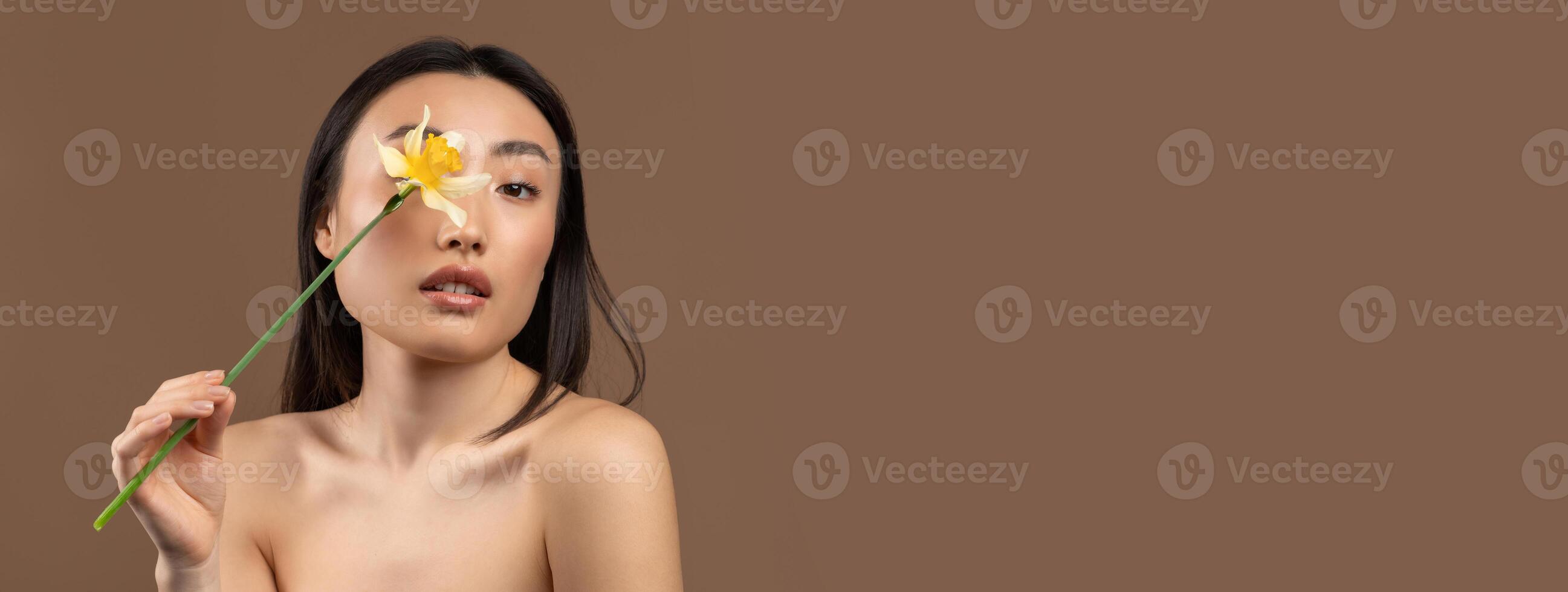 Tender korean lady holding beautiful daffodil flower and closing eye, posing over brown studio background and looking at camera. Portrait of asian woman having silky skin and nude makeup, copy space photo