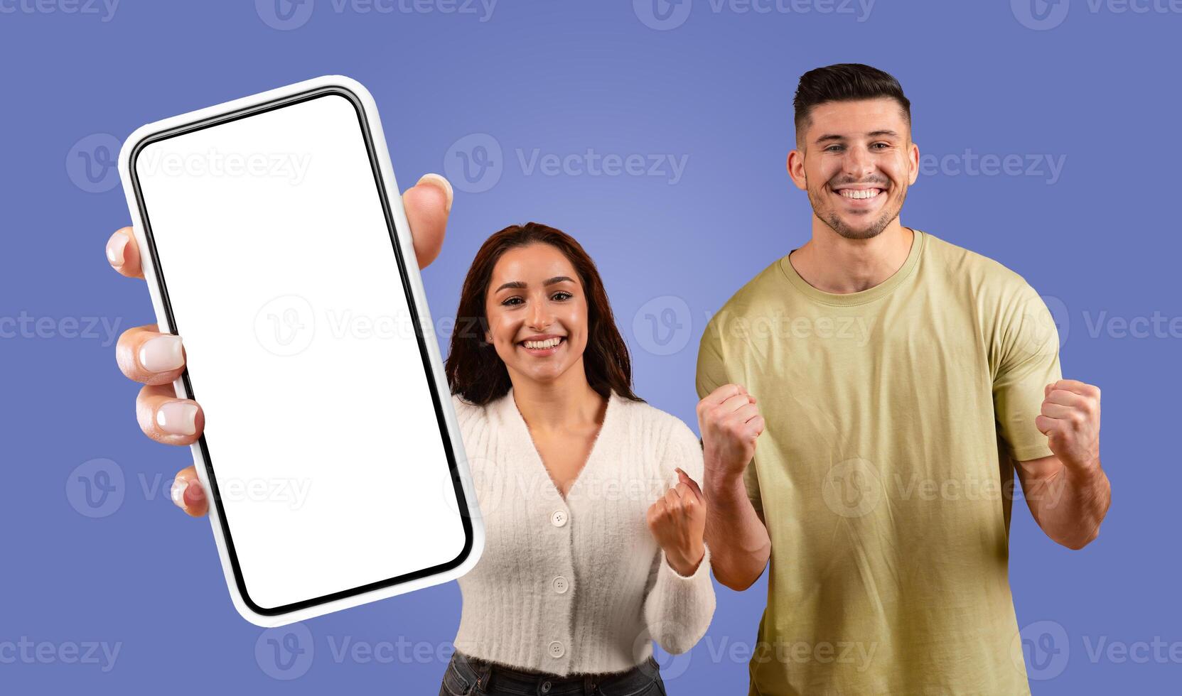 A young woman holds a smartphone with a white screen in front of her while smiling and celebrating with a man. The man has his fists clenched in the air and is also smiling. photo