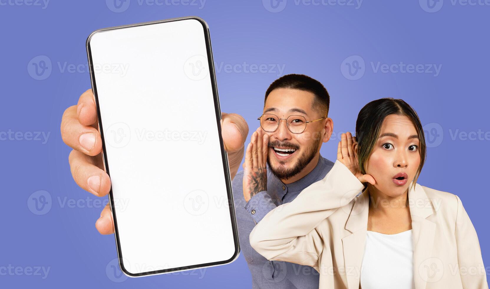 Asian man with a beard and glasses shouts into the screen of a smartphone held by an unseen hand. A woman with her hand to her ear listens intently, with a surprised expression on her face. photo