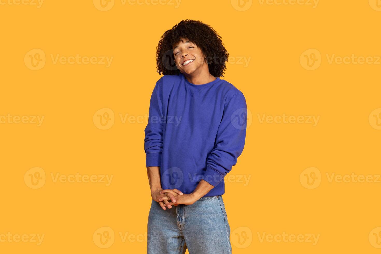 This image shows a young man smiling in front of a yellow background. He is wearing a blue sweatshirt and jeans. He has a happy and relaxed expression, and appears to be confident and at ease. photo