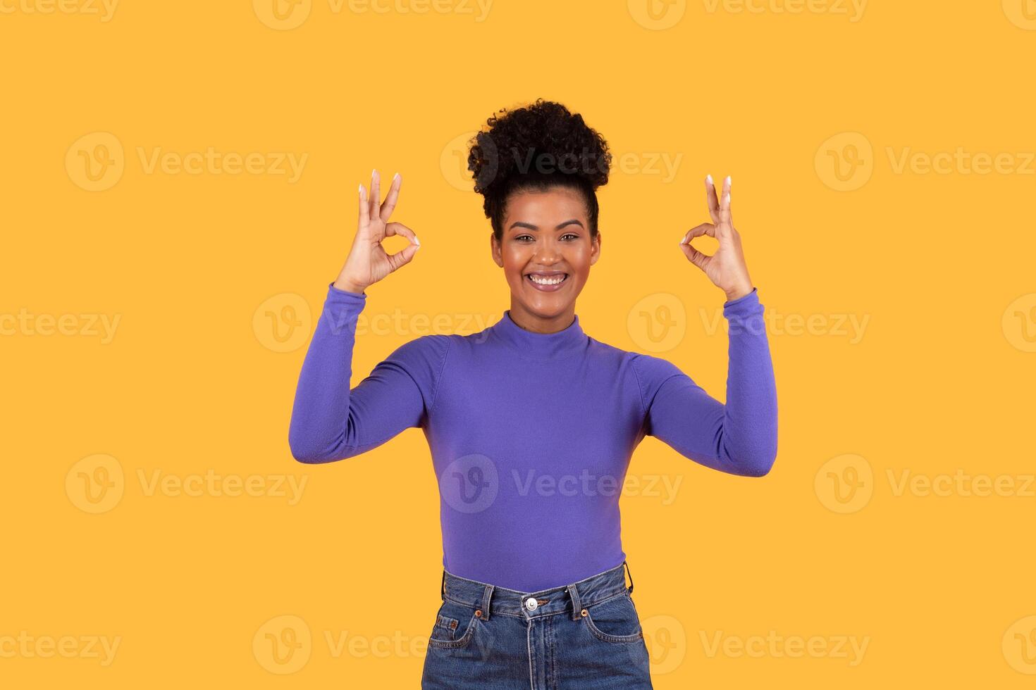 A smiling woman with an afro hairstyle is standing in front of a yellow background. She is wearing a purple long-sleeved shirt and blue jeans. She is making the okay sign with both hands photo