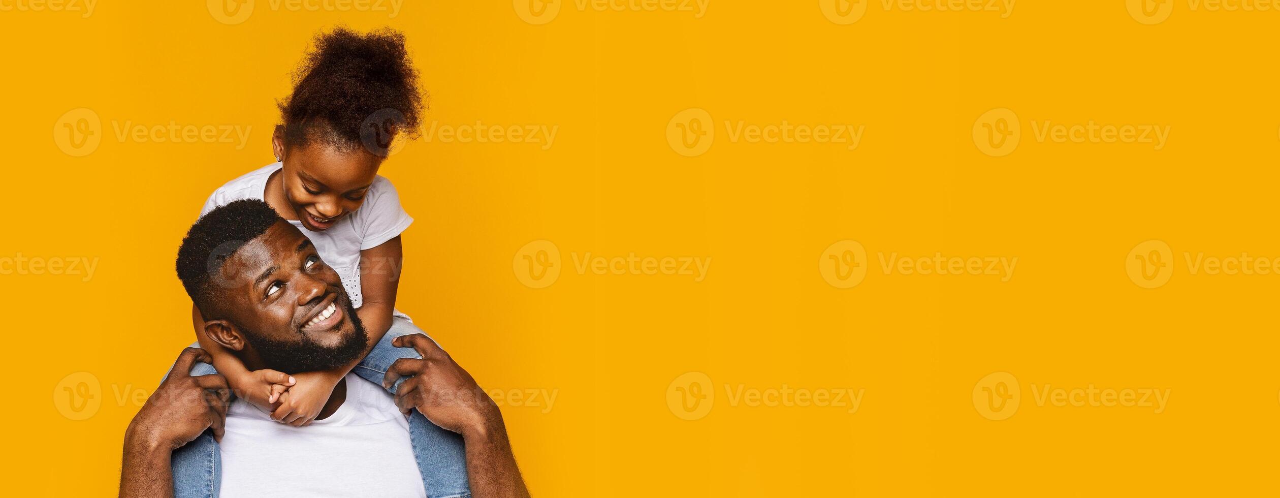 African American father is seated with a big smile while his daughter playfully rides on his back. The bright yellow background adds a vibrant and cheerful atmosphere to their connection photo