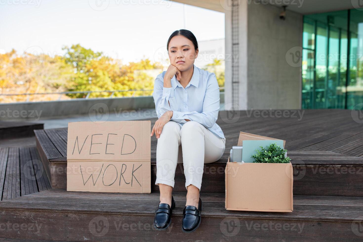 A young Asian woman is seated on the steps outside a modern building, looking pensive and concerned. She holds a sign that reads Need Work while a box filled with personal items photo