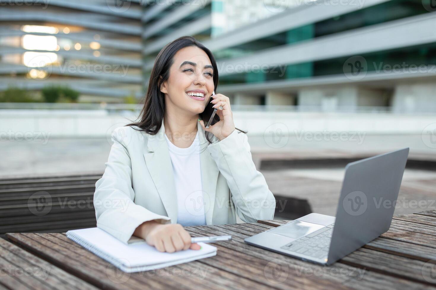 A young woman in a white blazer and white t-shirt sits at a wooden table outside a modern office building, smiling as she talks on her phone. A laptop is open in front of her, copy space photo