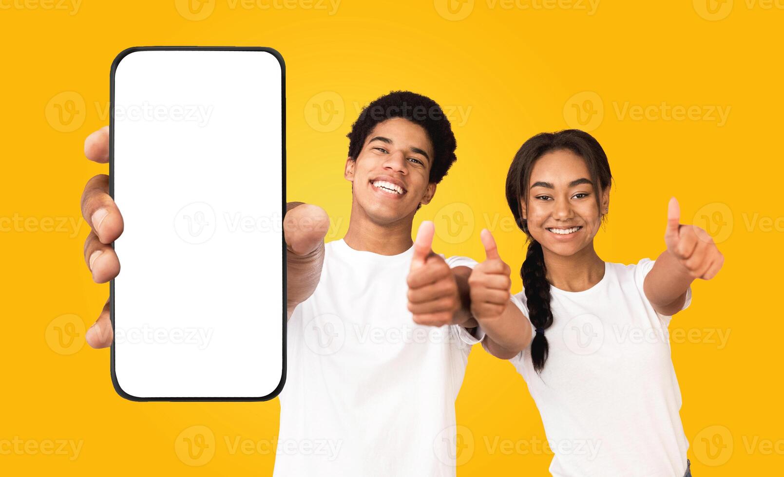 A young black man and woman are holding a smartphone with a blank screen. They are both smiling and giving thumbs up. The background is a solid yellow color. photo
