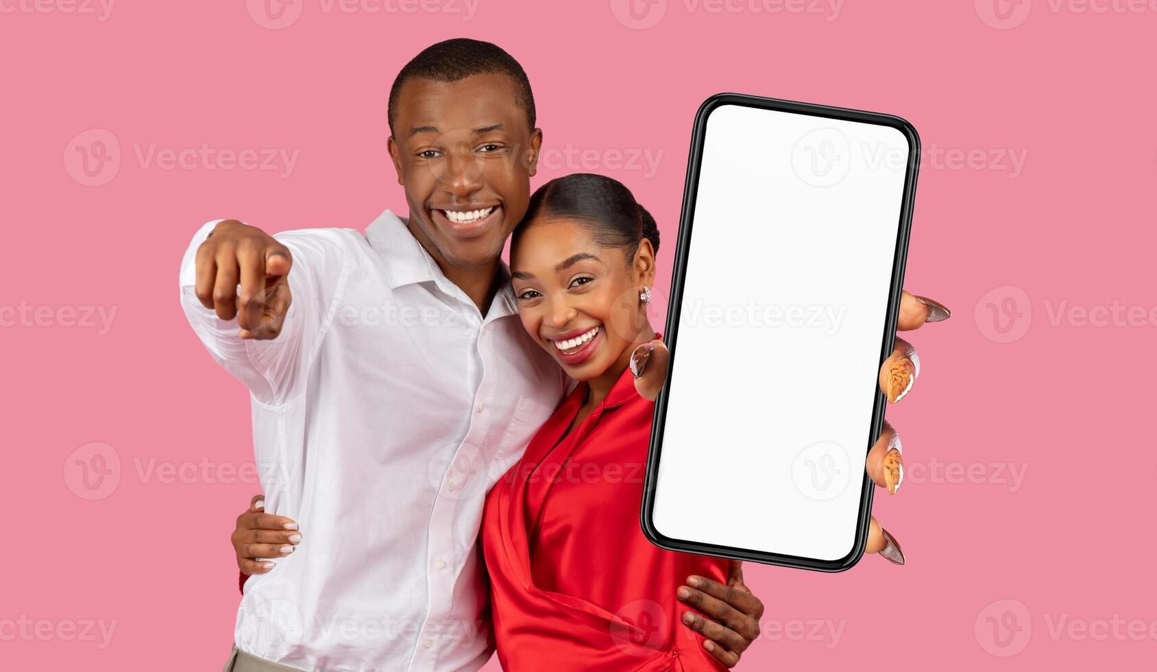 A happy black couple is standing in front of a pink background. The man is pointing towards the camera with his left hand. The woman is holding up a smartphone with a blank screen photo