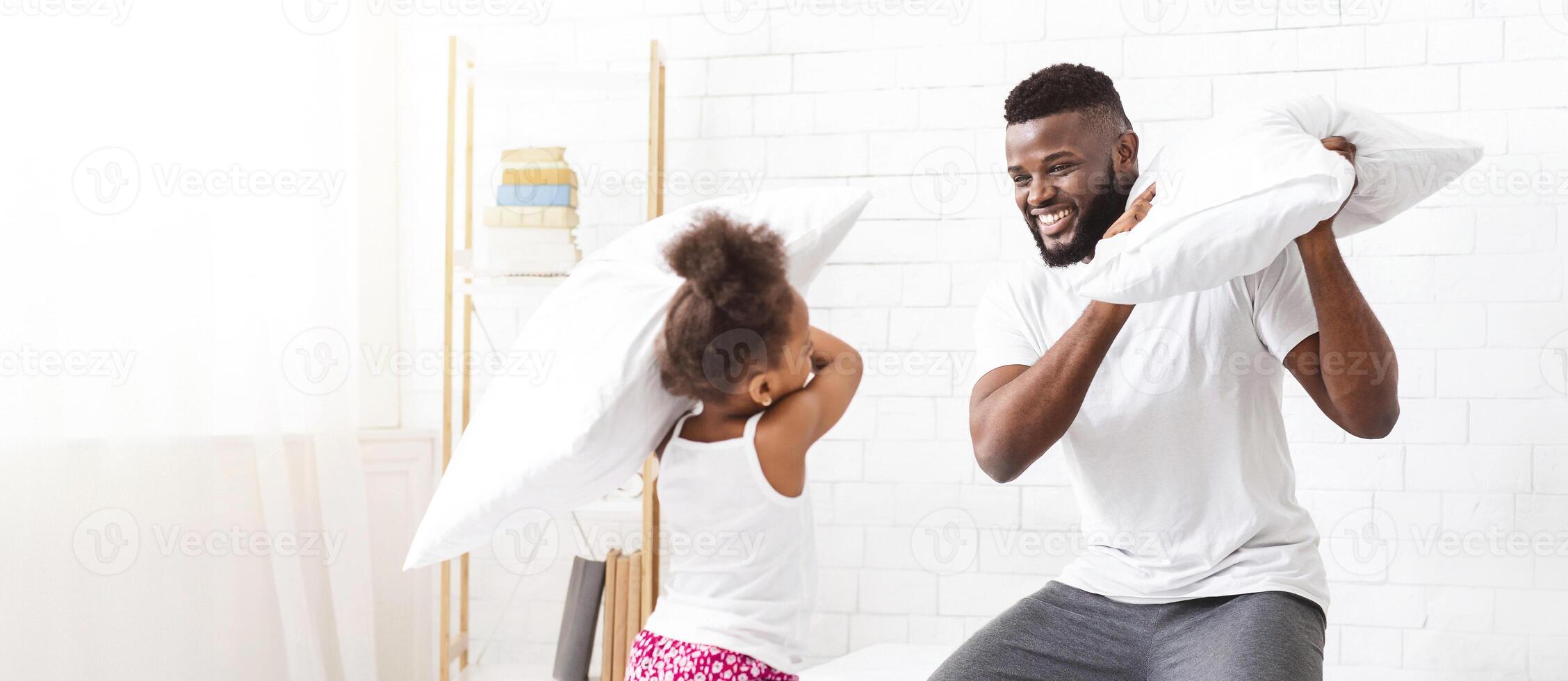 African American father and his daughter are having a fun pillow fight in their bright, white bedroom. The father is smiling as he holds up his pillow, while the daughter looks playful photo