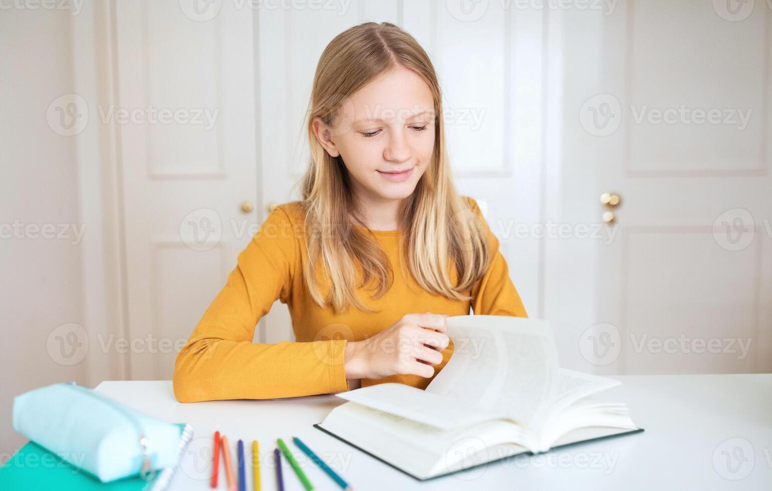 A teen girl with long blonde hair sits at a white desk and studies from a textbook. Her right hand rests on the open book as she looks down at the pages. photo
