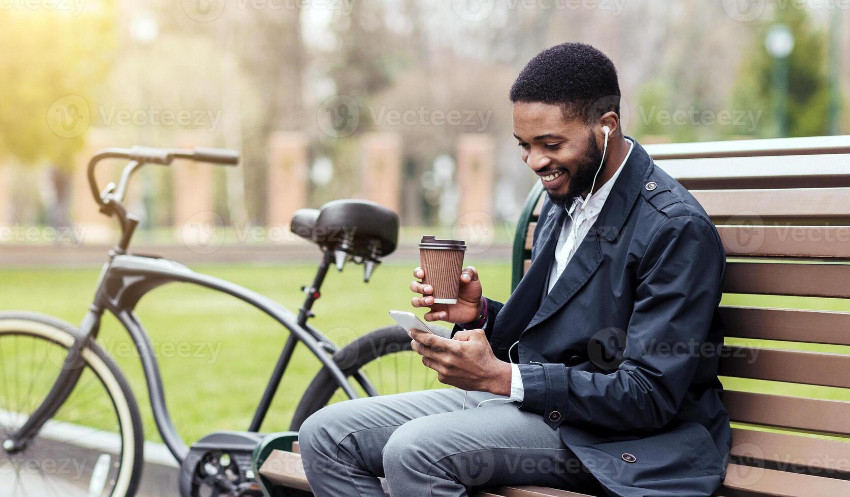 A professional black man is in a park using his phone, with his bicycle beside the bench. The scene is serene and urban, showing a balance between professional duties and leisure. photo