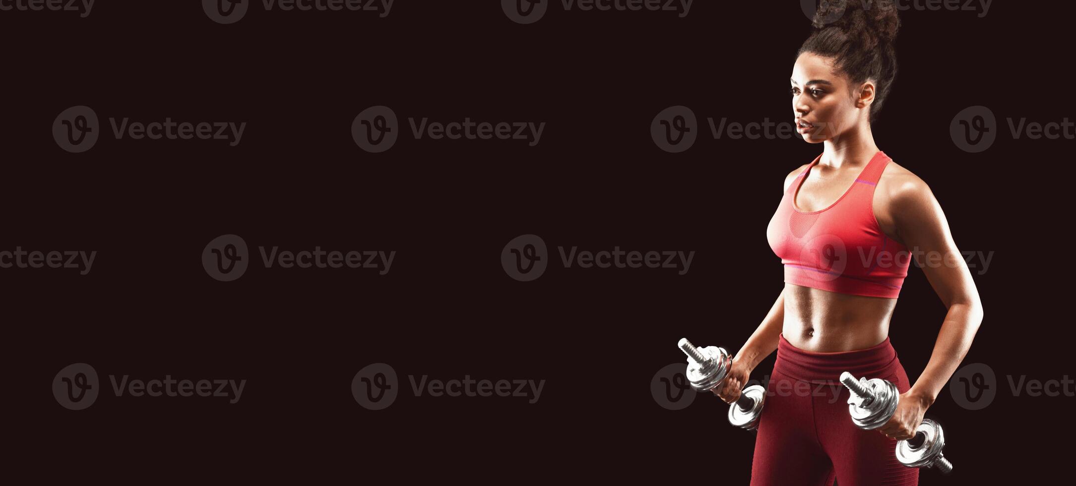 Concentrated black sporty girl holding barbells and looking at free space over black background photo