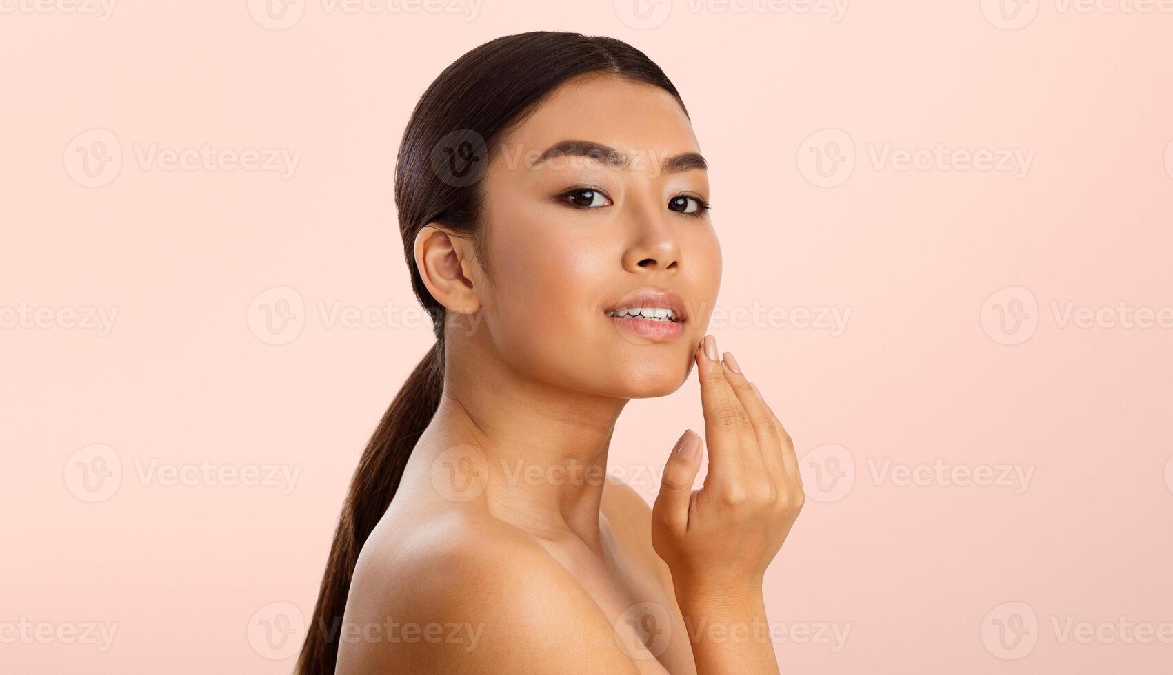 A young Asian woman with brown hair, wearing no makeup, is touching her cheek with her right hand. She is looking to the left, and her expression is relaxed and confident. photo