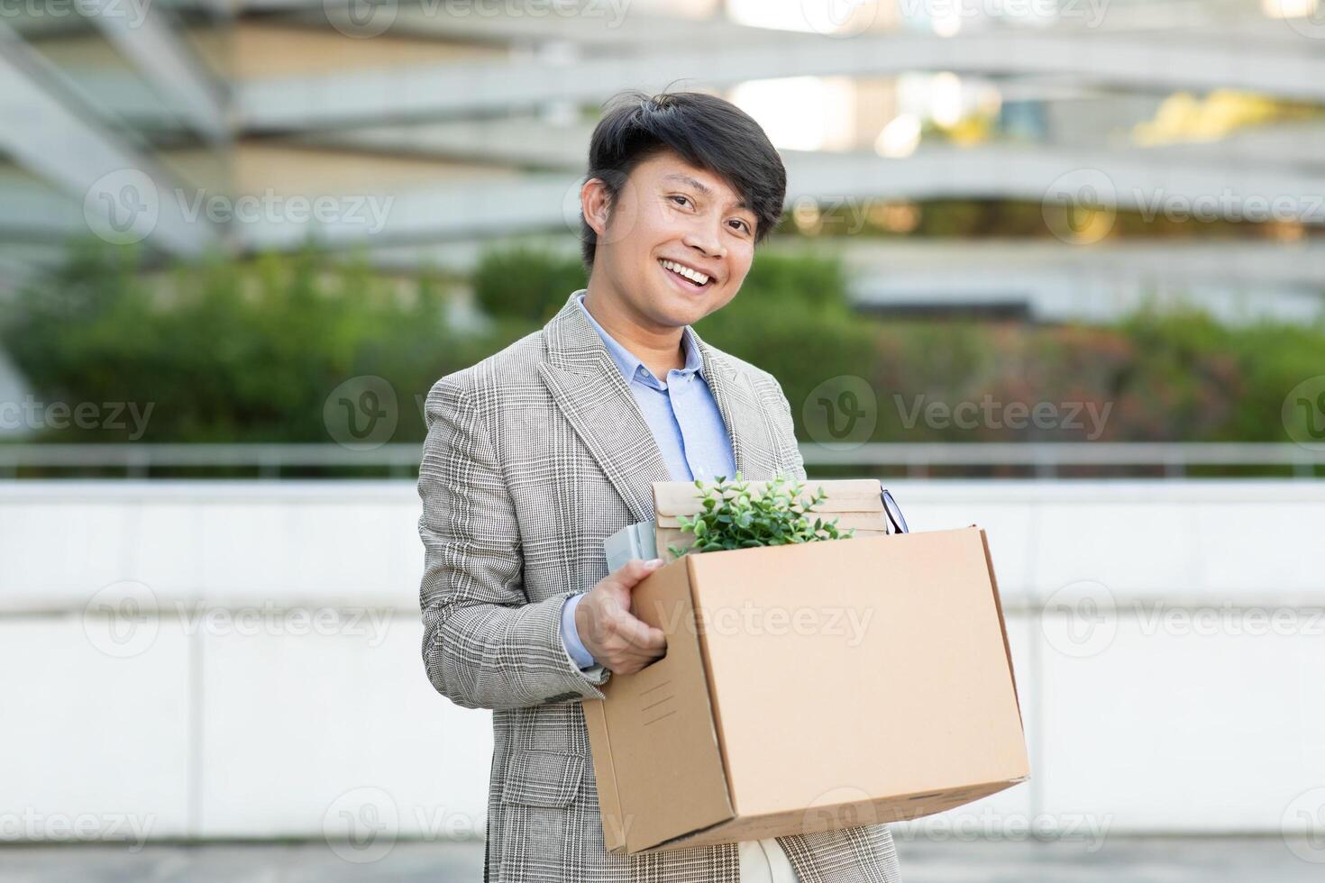 Asian man in a plaid blazer smiles as he carries a box full of personal belongings outside of a modern office building. photo