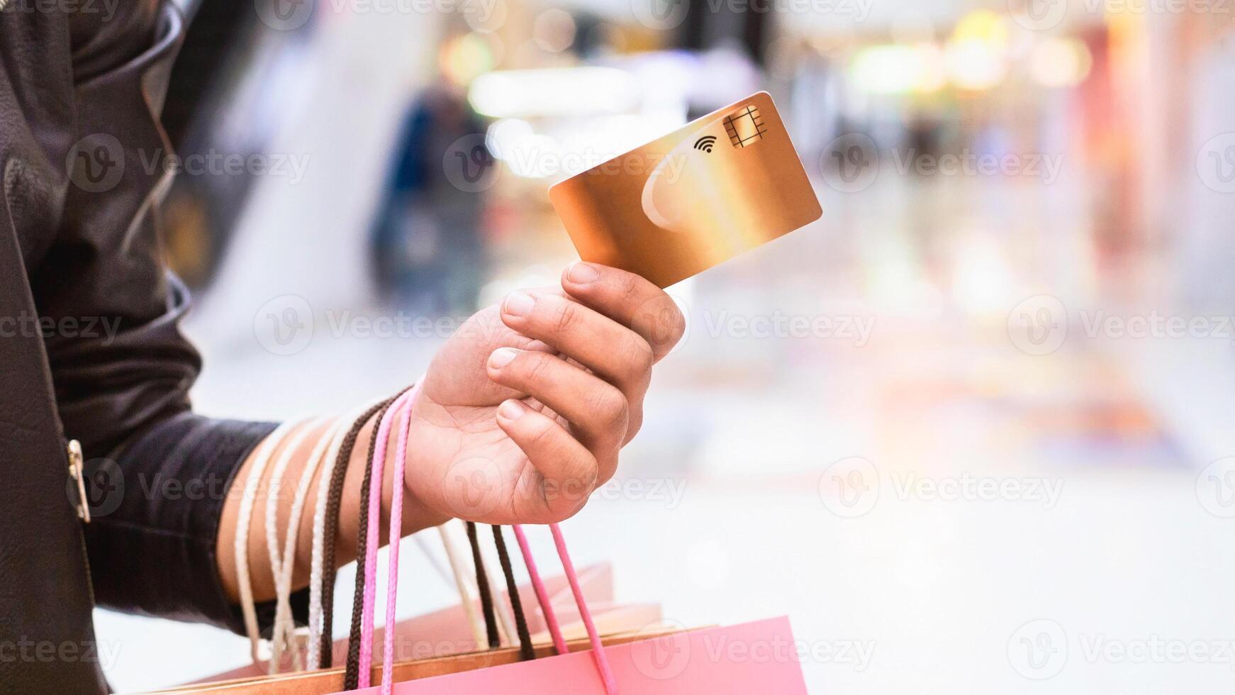 Unrecognizable woman is seen holding a credit card while shopping in a busy mall. She appears focused on making a purchase, surrounded by various stores and shoppers, cropped photo