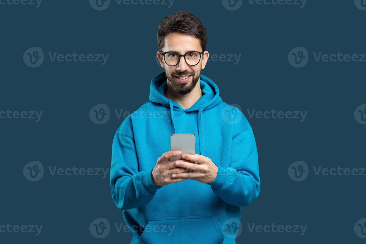A young man with glasses and a beard stands indoors, wearing a vibrant blue hoodie. He smiles brightly as he interacts with his smartphone, conveying a sense of joy and engagement. photo