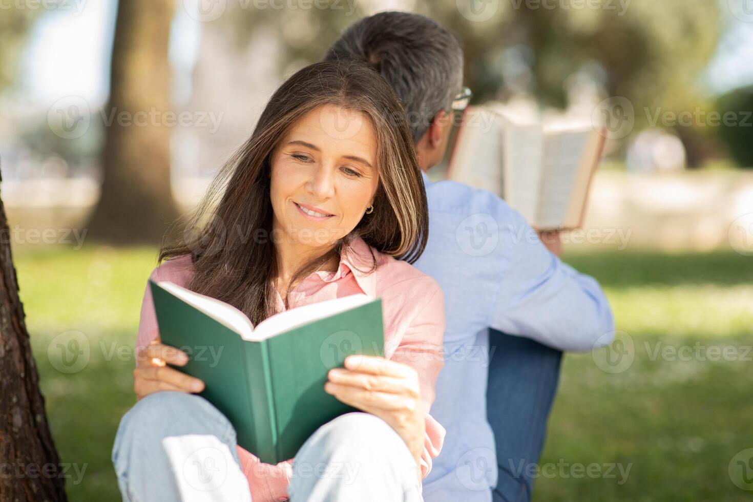 A woman enjoys a book while seated against a tree, her back to a man who is also reading. The warm sunlight filters through the branches, creating a peaceful atmosphere in the park. photo