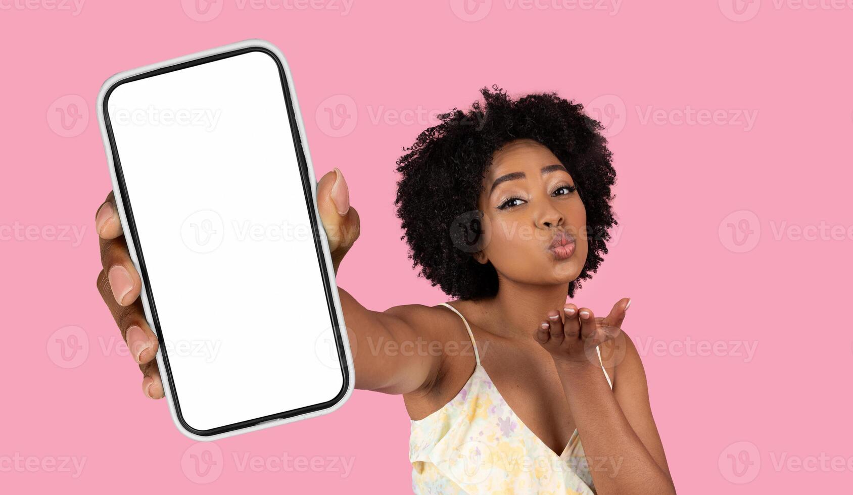 A young black woman with curly hair is holding a smartphone with a blank white screen in her outstretched hand. She is looking directly at the camera and blowing a kiss. photo