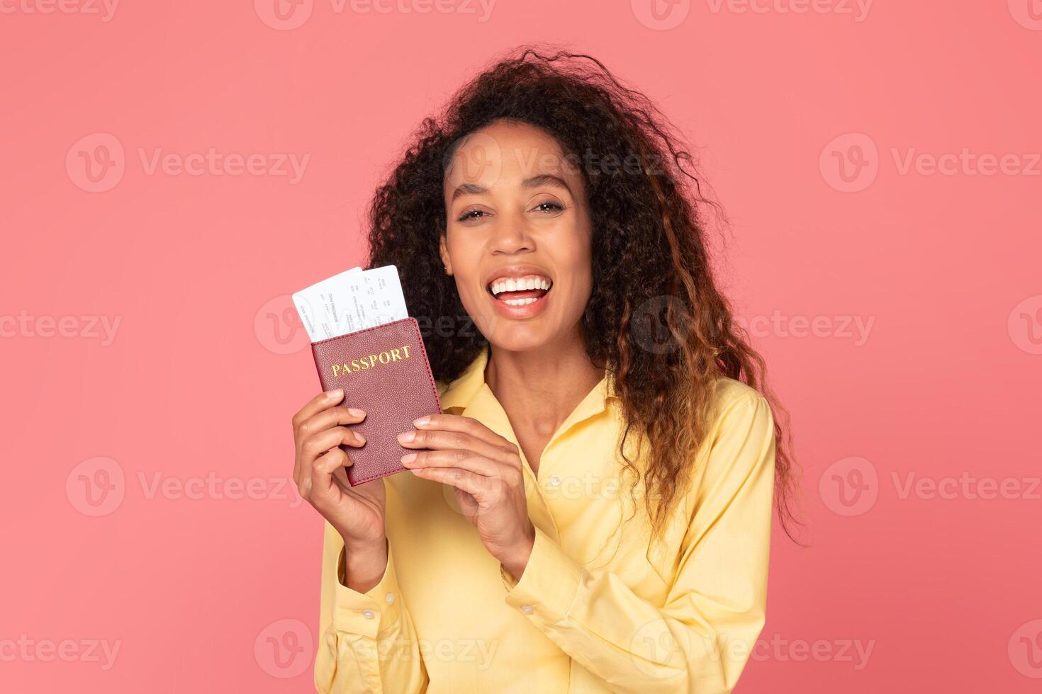 A young African American woman with curly hair is smiling and holding a passport and boarding pass in her hands against a pink background. photo