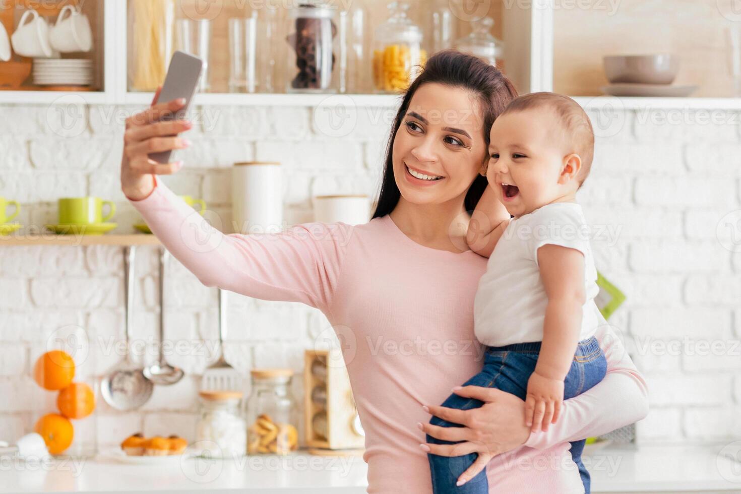 A woman holding her baby in her arms while taking a selfie with a smartphone. The mother and child are smiling at the camera in a casual and joyful moment. photo