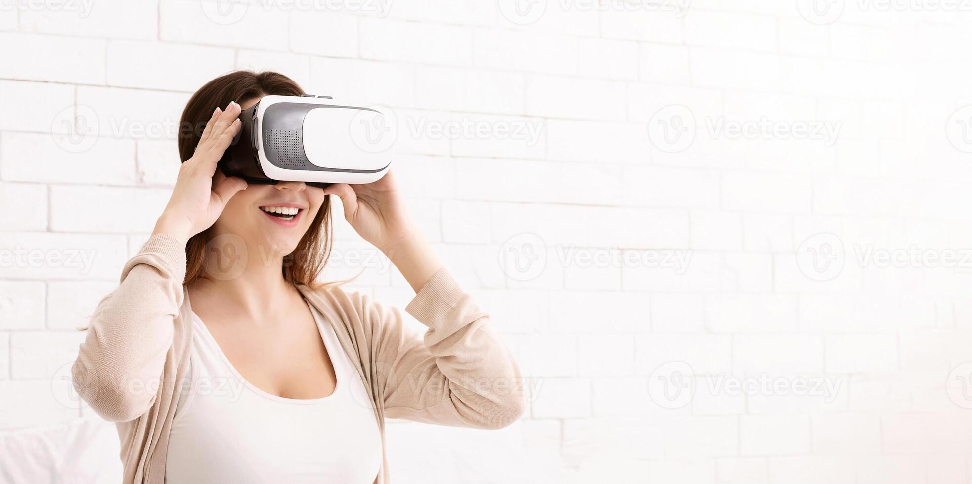 A young woman is using a virtual reality headset and smiling in a brightly lit room. She seems to be thoroughly enjoying the immersive digital experience. photo