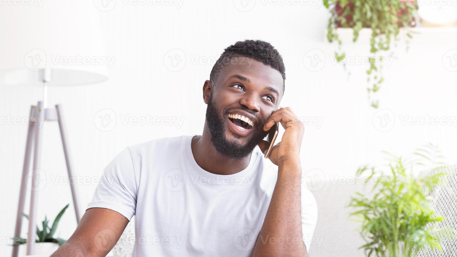 African American man with a beard smiles while talking on a phone in his home office. He is wearing a white shirt and sitting in front of a white wall with a floor lamp next to him. photo