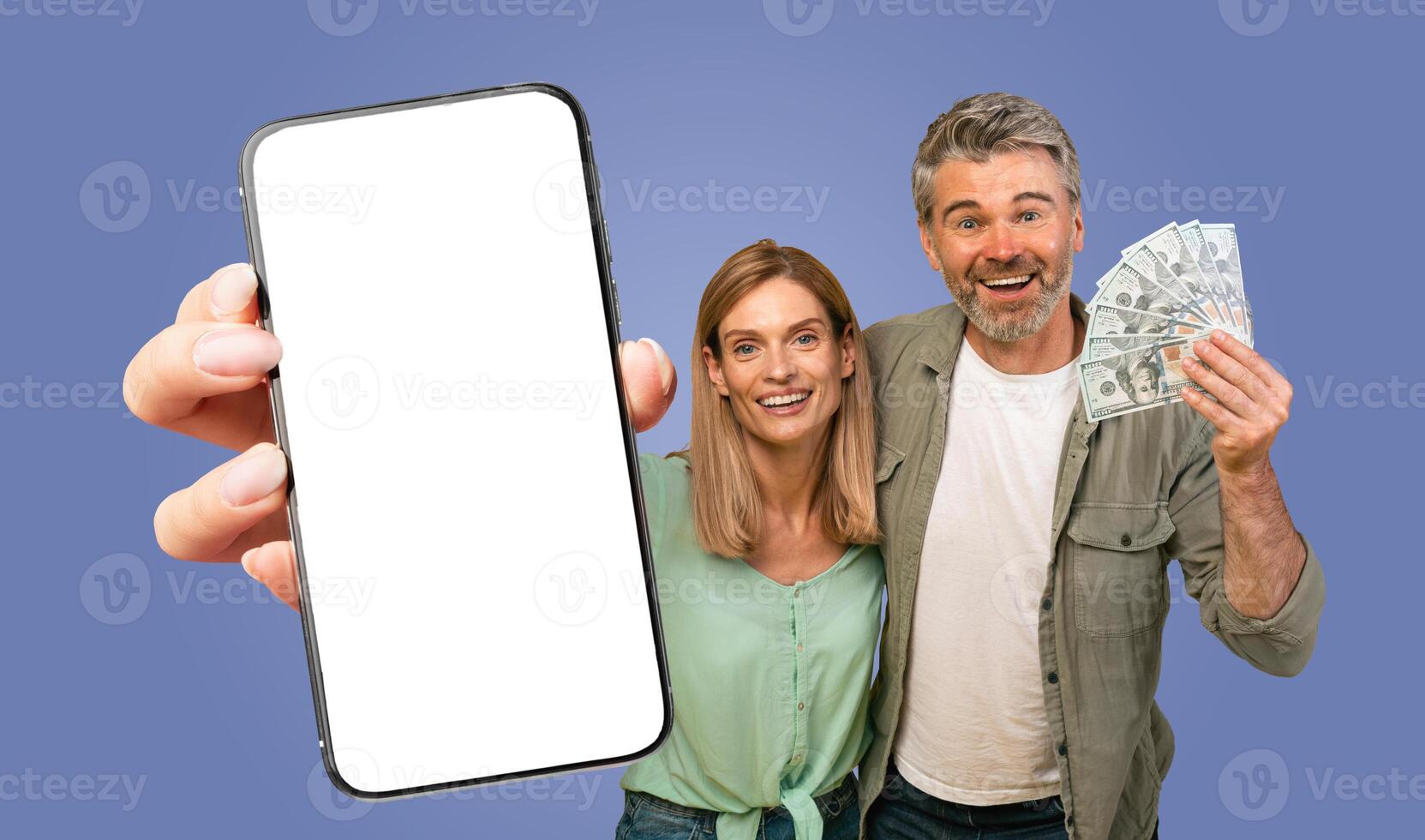 A couple is shown smiling and holding a smartphone with a blank screen and cash in front of a blue background. The woman is holding the smartphone while the man is holding a fan of cash. photo