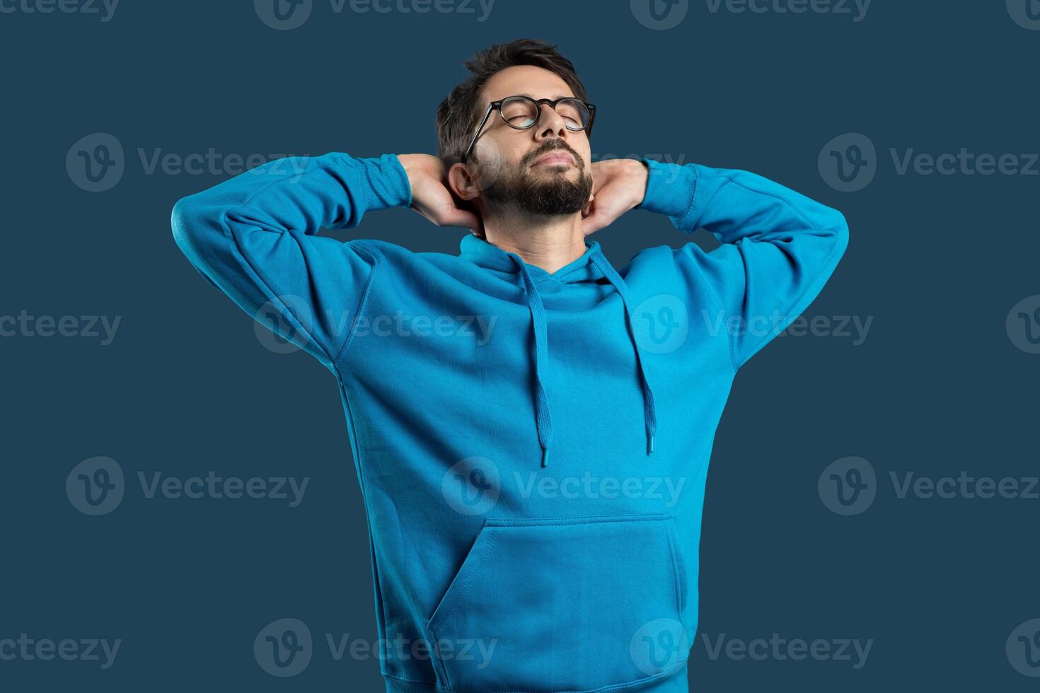 A young man wearing glasses and a casual blue hoodie stands with his hands behind his head, eyes closed in a gesture of relaxation and contentment, blue background photo