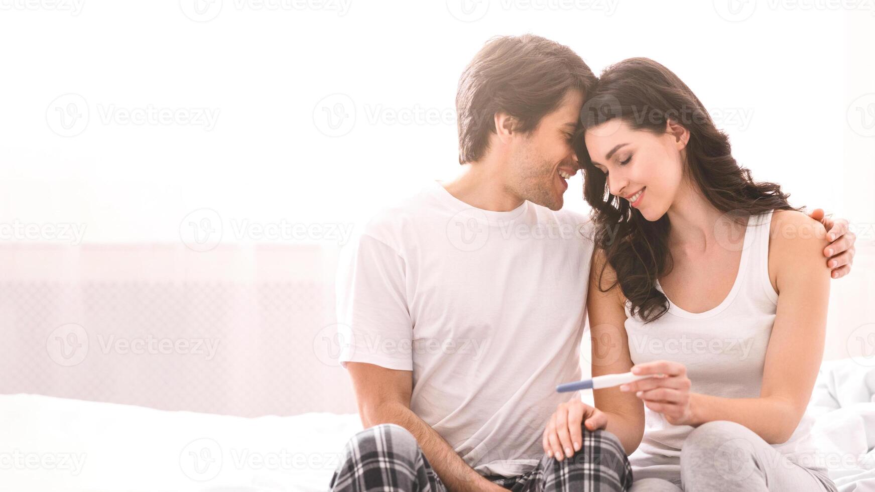 A couple is sitting on a bed looking at a pregnancy test, they both have smiles on their faces. The woman is holding the test and the man has his arm around her shoulder. photo
