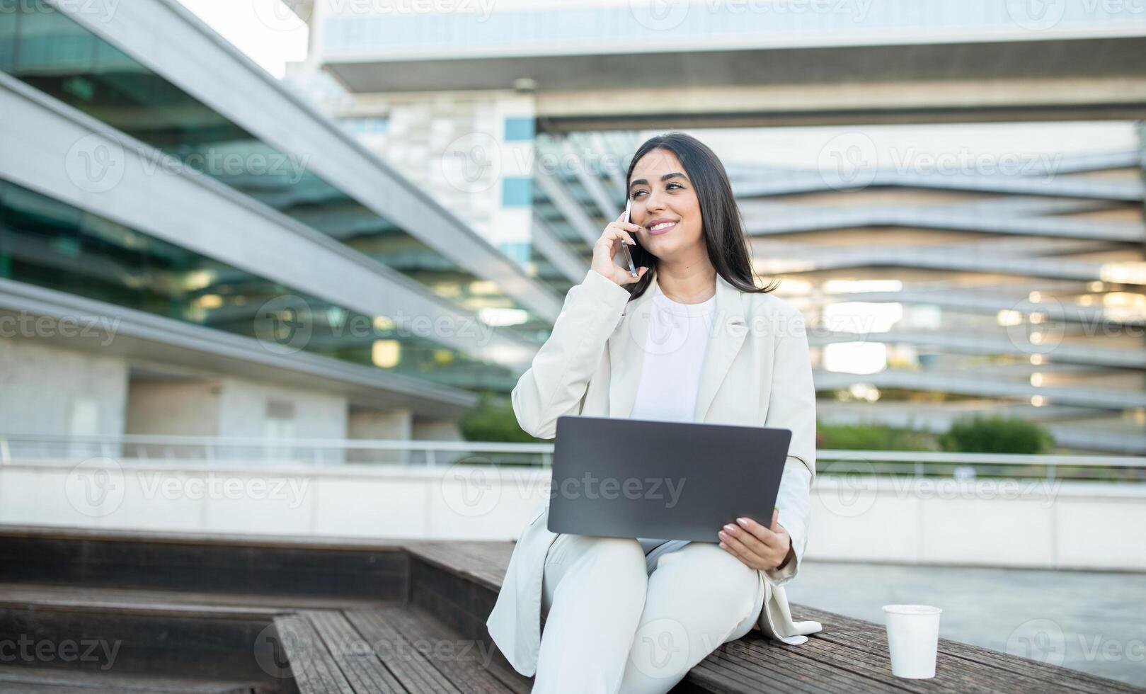 A young businesswoman is sitting on a bench in front of a modern office building while holding her laptop and talking on her phone. She is wearing a white blazer and pants. photo