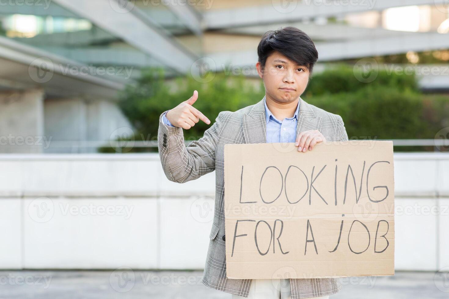 Asian young man stands outside in an urban setting, holding a cardboard sign that reads Looking for a Job while pointing at himself. He wears a light-colored jacket, white shirt, and white pants. photo