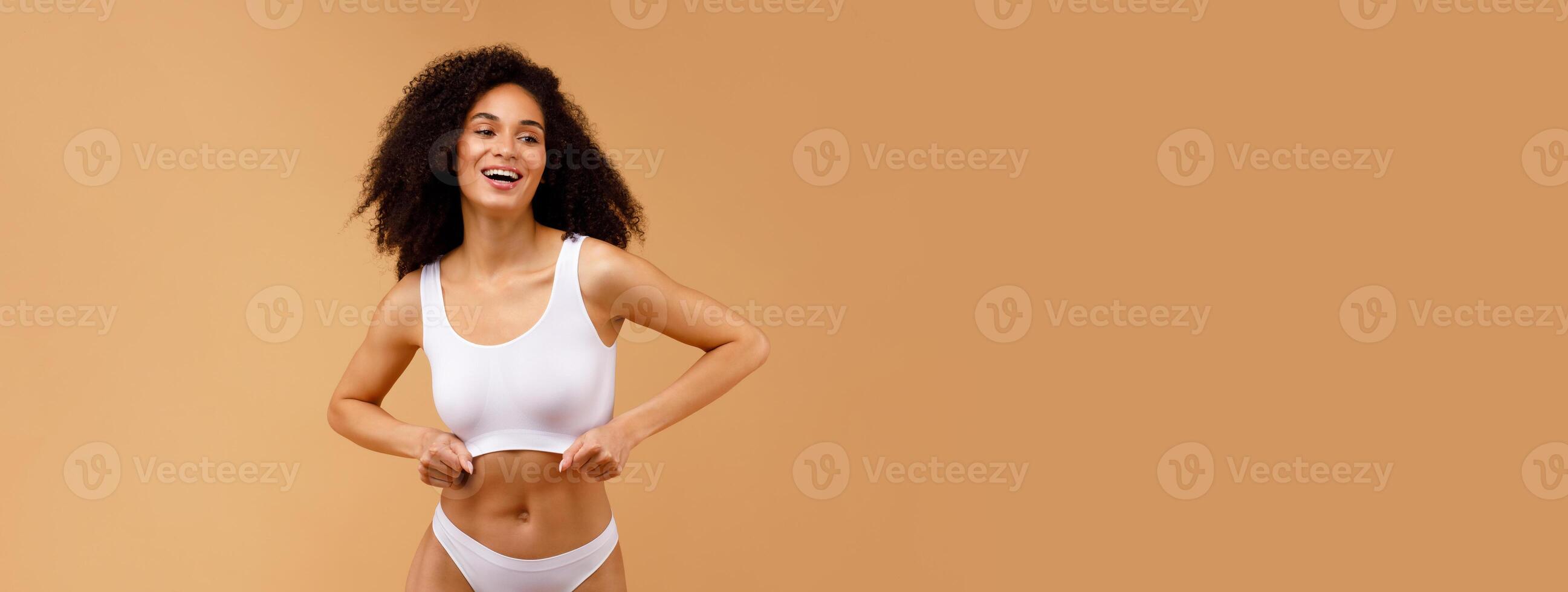 Slim african american woman in white underwear top bra and panties standing isolated over beige studio background, posing and smiling, free space photo