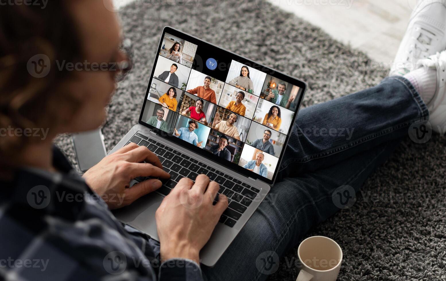 A man sits on the floor with his legs crossed, using a laptop to participate in a group call. The laptop screen shows a grid of multiple participants in the call. photo