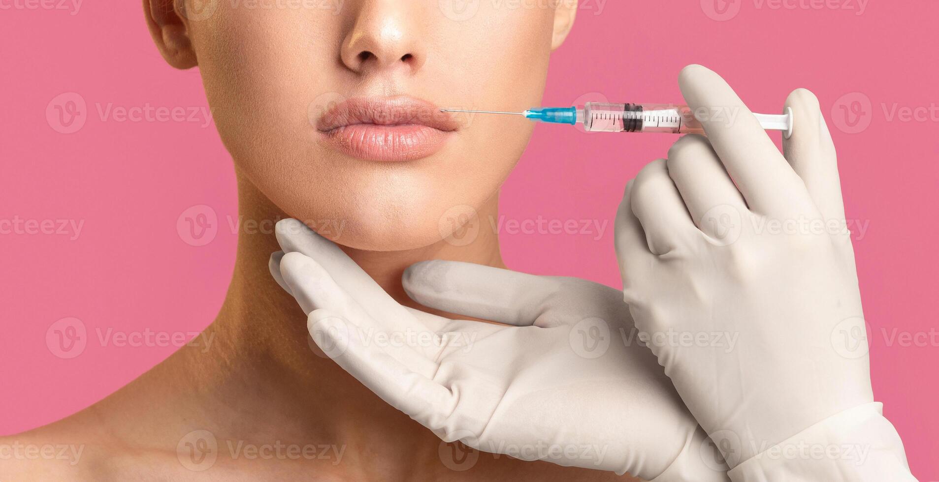 Cropped of woman receiving lip injections, focusing on her lips and the gloved hand holding the syringe. The woman is looking straight ahead, and the background is a solid pink color photo